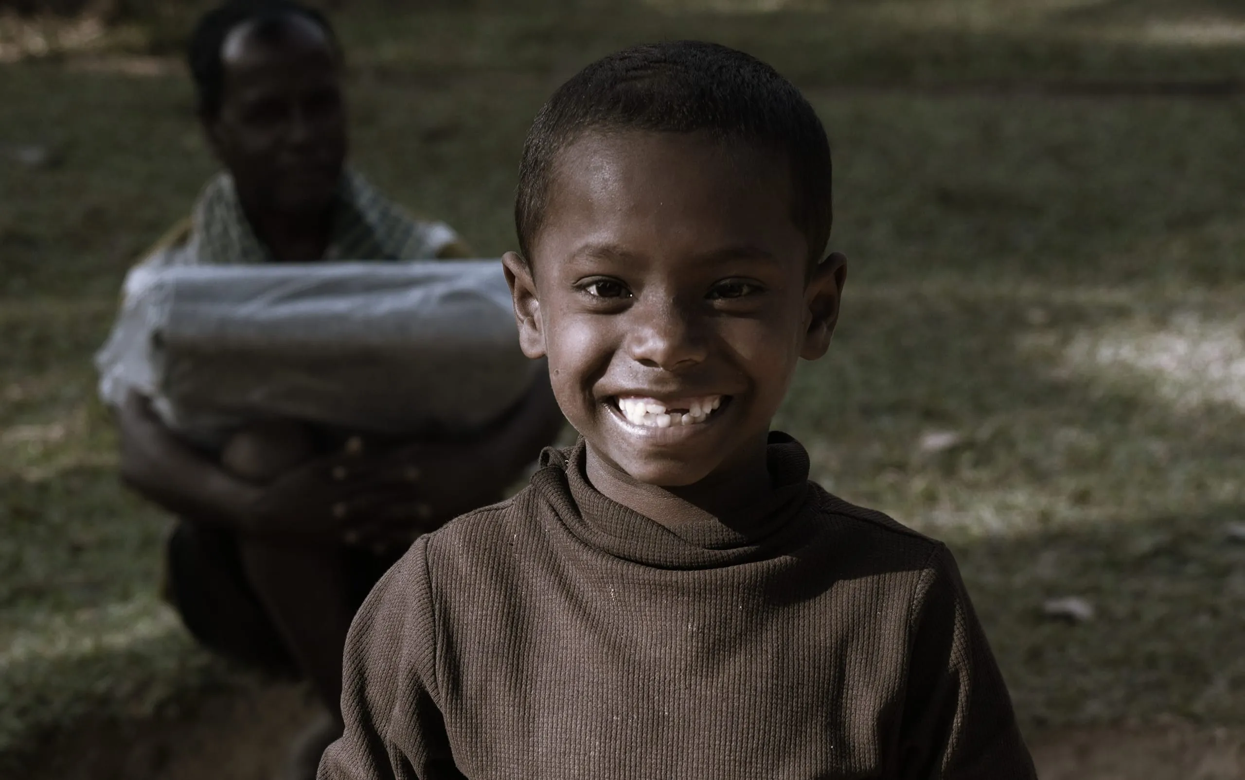 A young boy of the tea garden community grinning, a relief package being carried behind him