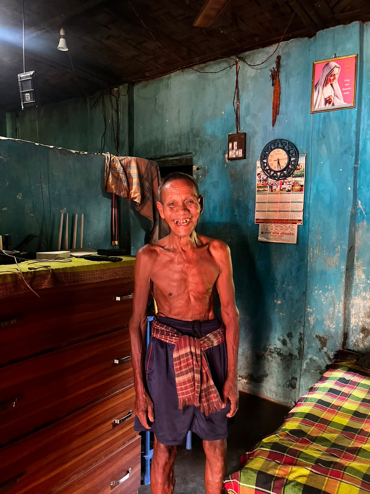 An elderly tea worker smiling inside his small home, his ribcage visible from years of malnutrition.