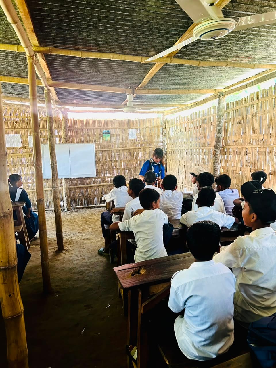 A HITH volunteer teaching students seated on wooden benches inside a bamboo-walled classroom.